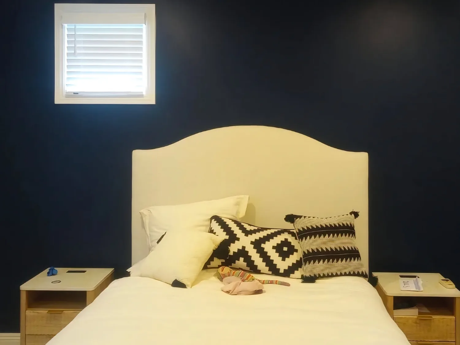 Bedroom wall painted a deep navy behind a cream upholstered headboard, with a white-framed shuttered window to the left and pale wood nightstands either side of the bed