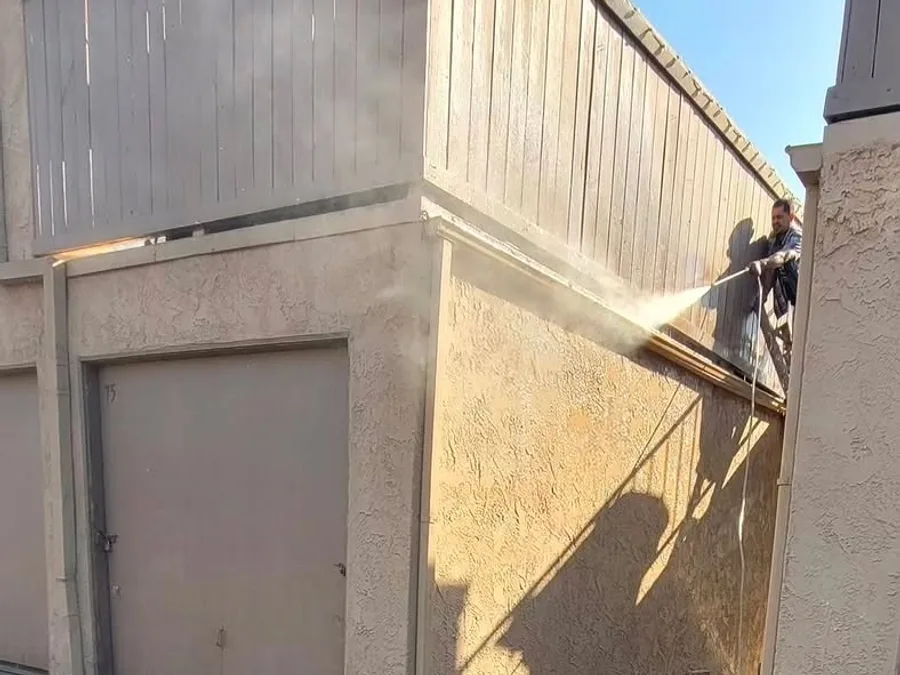 One of the crew washing the gutters down in the breezeway of a condo building, spray coming off the wall in bright sun