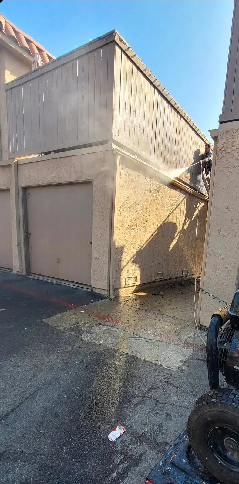 One of the crew in the breezeway of a condo building, washing the gutters and roof line down with a pressure washer, spray coming off the wall in bright sun