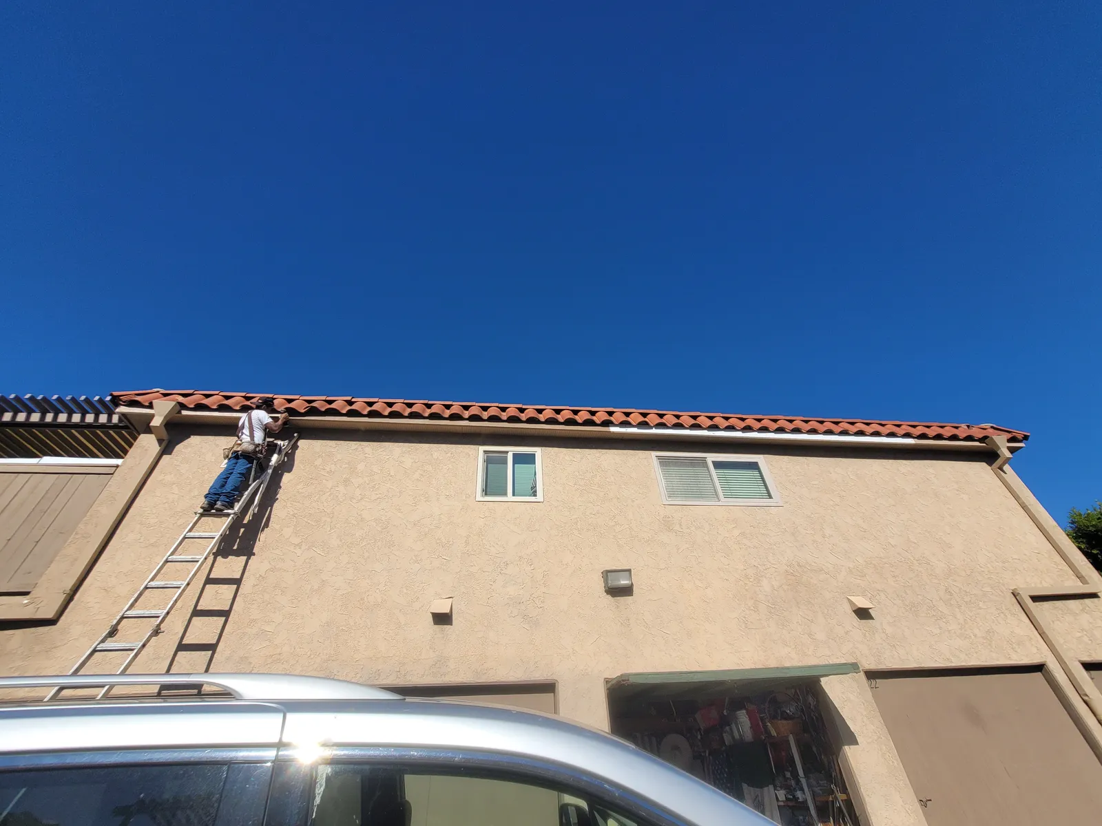 An extension ladder set against a two-story stucco building with a tile roof, one of the crew working on the fascia at the roof line under a clear blue sky