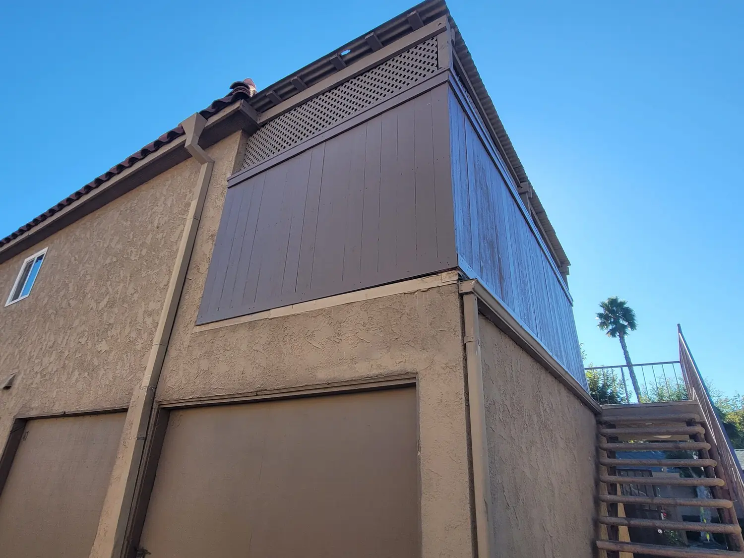 Two-story stucco building shot from below, with a wood board and lattice screen wall whose left panel is freshly painted brown and right panel is still weathered grey